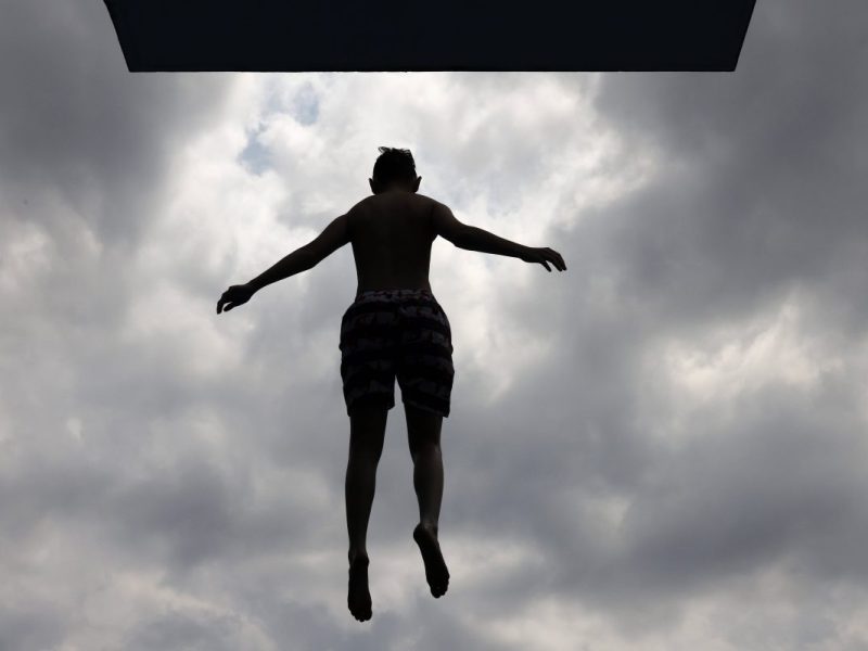 Perspektive von unten, Kind springt von Brett ins Wasser, grauer, wolkenverhangener Himmel