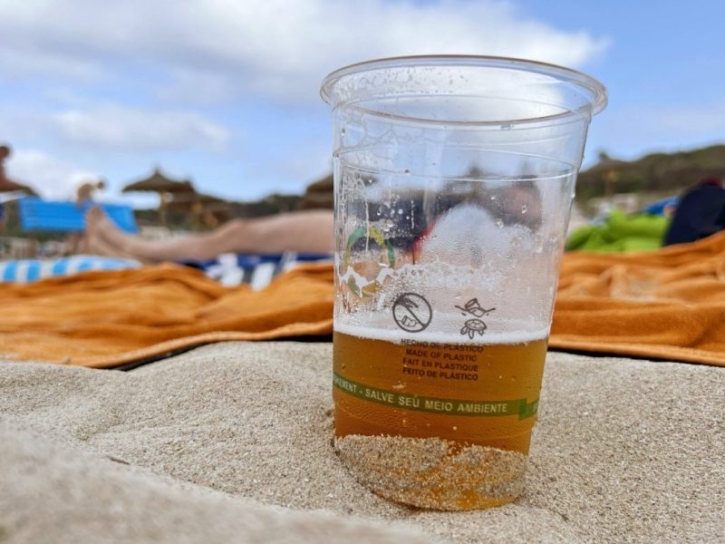 halbgefüllter Bierbecher am Strand von Mallorca, Urlauber auf Handtüchern im Hintergrund