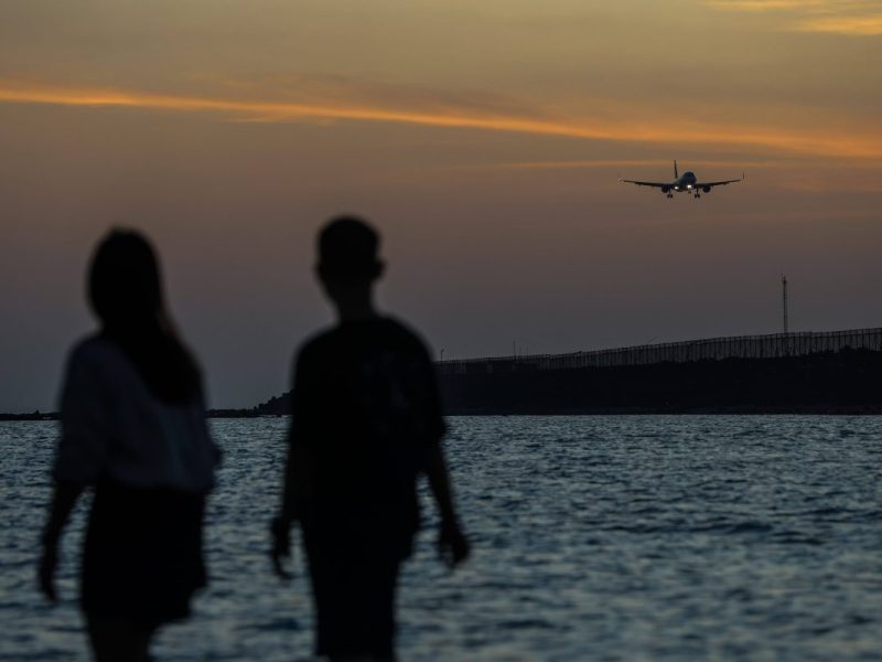 Touristen am Strand von Bali vor einem Sonnenuntergang und einem tieffliegenden Flugzeug