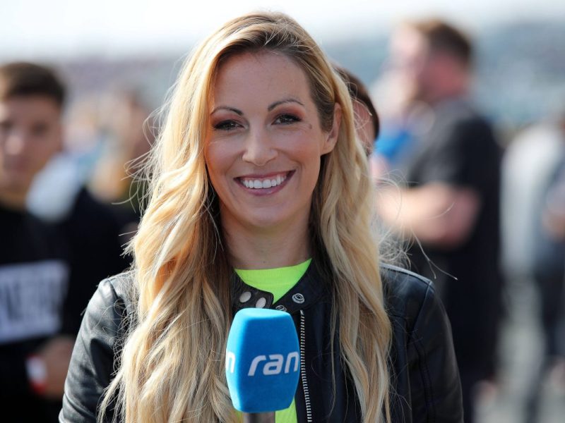 Andrea Kaiser steht mit einem Mikrofon in der Hand an einer Rennstrecke.
