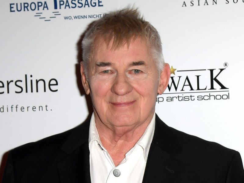 Schauspieler Heinz Hoenig. Er trägt ein schwarzes Sakko mit einem weißen Hemd drunter. Im Hintergrund ist eine weiße Wand.