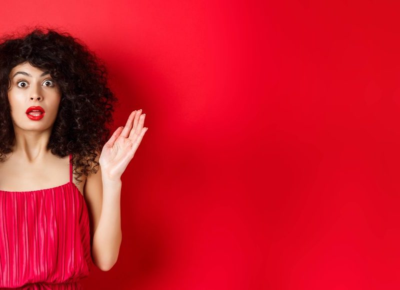 Das Bild hat einen roten Hintergrund, vor dem eine Frau steht. Die Frau trägt roten Lippenstift und ein rotes Kleid. Sie hat in ihrer linken Hand einen Lippenstift und schwarze lockige Haare. Sie hat ihren Mund auf und schaut erschrocken.